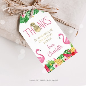 以下が含まれることがあります： ピンクのフラミンゴのサンキュータグ。金色のパイナップルと熱帯の葉が描かれています。テキストは「私の最初の誕生日を祝ってくれてありがとう！愛を込めて、シャーロット」です。