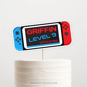 以下が含まれることがあります： 「GRIFFIN LEVEL 9 UNLOCKED」というテキストが書かれた、青と赤の任天堂スイッチゲームコンソールケーキトッパー。