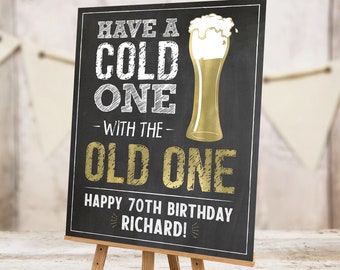 Può includere: Un cartello nero in ardesia con scritta in gesso bianco che recita "Have a Cold One - With the Old One Happy 70th Birthday Richard!" Un bicchiere di birra dorato con schiuma bianca è a destra del testo. Il cartello è su un cavalletto di legno.