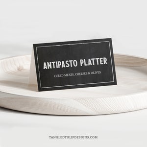 Könnte beinhalten: Ein schwarzes Schild aus Schiefer mit weißem Text, der "Antipasto Platter Cured Meats, Cheeses & Olives" lautet. Das Schild steht auf einem weißen Holztablett.