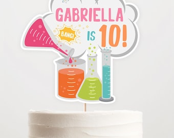 May include: A cake topper featuring a white cloud with the text "Gabriella is 10!" in pink, orange, and gray. The cloud has a small star and a speech bubble with the word "Bang!" in it. There are also four beakers and a pink funnel pouring liquid into a beaker.