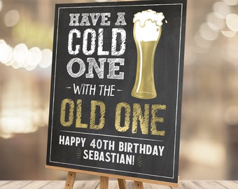 Può includere: Un cartello in ardesia nera con lettere bianche e dorate. Il cartello dice "Have a cold one with the old one. Happy 40th Birthday Sebastian!"  Il cartello ha una grafica di bicchiere di birra dorato con un top di schiuma bianca.
