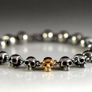 May include: A silver skull bracelet with a gold skull charm in the center. The bracelet is made of small, interconnected skull beads.