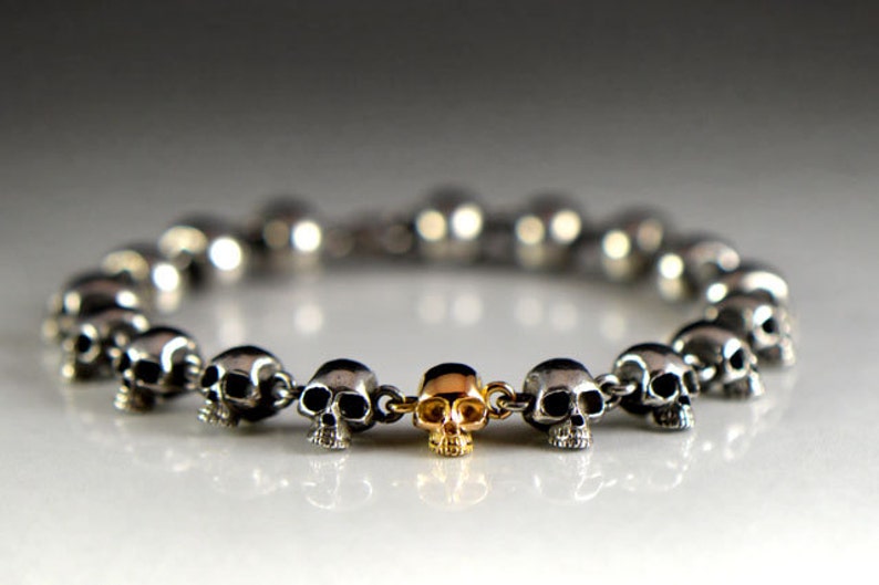 May include: A silver skull bracelet with a gold skull charm in the center. The bracelet is made of small, interconnected skull beads.