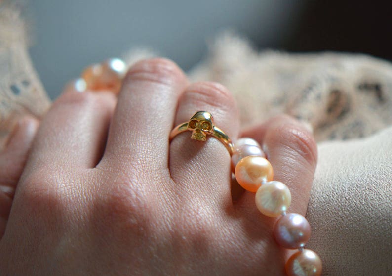 May include: A gold ring with a skull design on a finger wearing a pearl necklace.