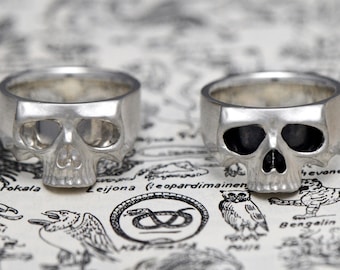 May include: Two silver skull rings. One ring has a solid skull design, while the other has a skull design with black enamel eyes.