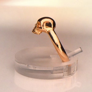 May include: A gold skull ring with a detailed design. The ring is displayed on a clear plastic ring holder.