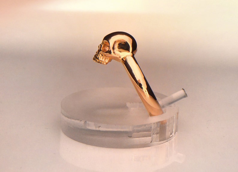 May include: A gold skull ring with a detailed design. The ring is displayed on a clear plastic ring holder.