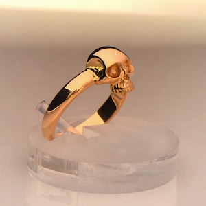 May include: A gold skull ring with a detailed design. The ring is on a clear display stand.
