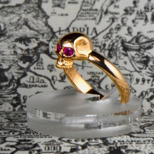 May include: A gold skull ring with ruby red eyes. The ring is on a clear display stand.