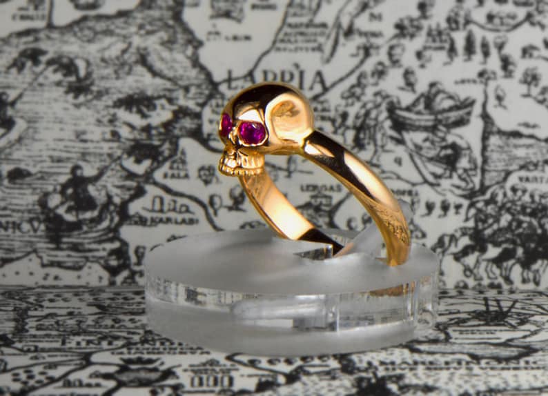 May include: A gold skull ring with ruby red eyes. The ring is on a clear display stand.