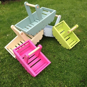 May include: A group of brightly painted wooden baskets in various colours, including pink, green, blue, and cream. Each basket has a wooden handle. The baskets are rectangular with slatted sides, set on green grass.