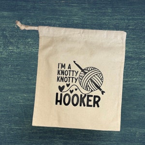 May include: A cream-colored drawstring bag with the text "I'M A Knotty Knotty Hooker" in black, along with a graphic of a yarn ball and crochet hook. The bag is made of a natural fabric.