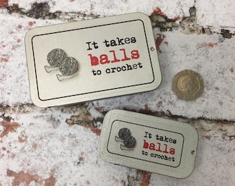 May include: Two rectangular silver tins with the text "It takes balls to crochet" in black and red. The tins feature a graphic of two yarn balls. A coin is next to the tins.