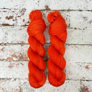 Könnte beinhalten: Zwei Knäuel orangefarbenes Garn, jeweils zu einer Kugel geformt. Das Garn ist in einem leuchtenden, kräftigen Orange.