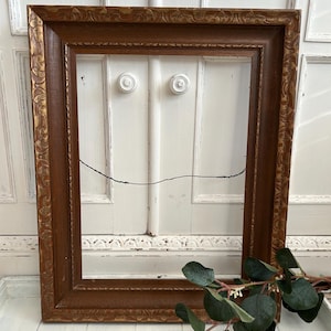 May include: A rectangular wooden picture frame featuring a dark brown finish with gold-toned decorative carved detailing along the inner and outer edges. A thin black wire is attached to the back for hanging purposes, and the frame is empty.