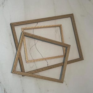 May include: Three gold-coloured picture frames of different sizes are arranged on a white surface. The frames have a decorative, ridged edge. Some have visible wire for hanging. The frames are empty, with no artwork or photos inside.