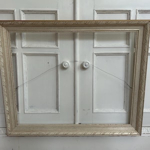 May include: A rectangular picture frame with a light beige, weathered finish and ornate, carved detailing along the inner and outer edges. The frame features a thin metal hanging wire attached to two circular white mounting points on the back.