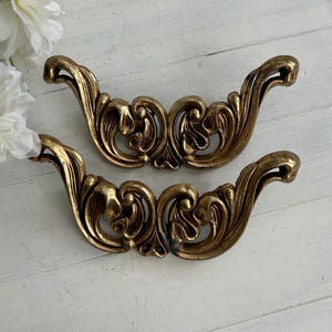 May include: Two ornate, gold-colored drawer pulls with a scrolling, floral design. The pulls have a curved shape with detailed embellishments, perfect for adding a vintage touch to furniture. The hardware is set against a white, distressed wood background.