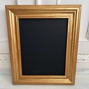 May include: A rectangular picture frame with a metallic gold finish and a stepped, tiered profile. The frame features a smooth, flat black centre panel, providing a high-contrast surface. The design is symmetrical with clean, sharp edges throughout.