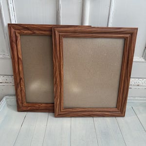 May include: Two rectangular picture frames featuring a medium brown wood grain finish with a textured, wavy pattern. Each frame has a wide, bevelled border and a neutral-coloured backing board visible within the centre opening of the frame.