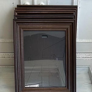 May include: A stack of rectangular dark brown wooden picture frames with tiered, stepped edges. Each frame features a clear glass front panel with a visible protective paper insert containing black line drawings of frame components and hardware.