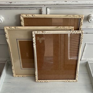 May include: Three off-white picture frames with ornate, carved edges. The frames are rectangular and vary in size, with a light brown backing visible through the glass. The frames are arranged on a light blue surface.