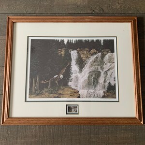 May include: Framed print of a bald eagle flying over a waterfall in a woodland setting. The print includes a small stamp of the eagle in the bottom right corner.