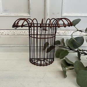 Peut inclure: Un panier décoratif cylindrique en fil de fer avec une finition marron rouille. Le panier a un dessus évasé avec des détails en fil d'arc et des supports verticaux en fil. Le panier est vide et repose sur une surface en bois blanc.
