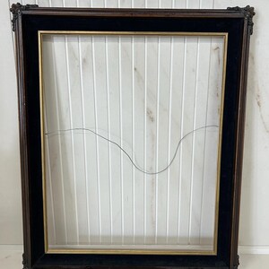 Peut inclure: Un cadre rectangulaire doté d'un bord extérieur en bois brun foncé, d'une bordure intérieure en velours noir et d'une fine garniture dorée. Le cadre comprend un fil de suspension métallique fin tendu horizontalement au centre de l'espace vide.