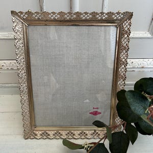 Peut inclure: Un cadre photo rectangulaire en métal doré avec une bordure ornée en filigrane. Le cadre contient un insert en tissu gris clair avec une étiquette rouge dans le coin inférieur droit sur laquelle est inscrit 8x10.