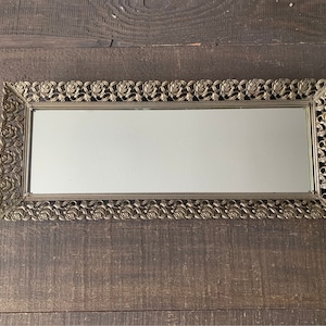 May include: A rectangular vanity mirror with a decorative gold-toned frame. The frame features a repeating rose pattern. The mirror is set against a dark wood surface.