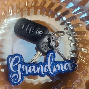 May include: A black car key fob and key with a blue glitter "Grandma" keyring. The keyring has white cursive lettering. The key fob has "Lock" and "Unlock" buttons.