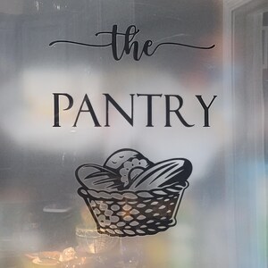 Op de afbeelding: Een matglazen deur met een zwarte vinyl sticker waarop "the PANTRY" staat, met daaronder een gestileerde broodmand.
