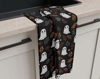 Könnte beinhalten: Schwarzes Küchentuch mit einem sich wiederholenden Muster aus weißen Geistern, orangefarbenen und braunen Blättern und kleinen weißen Blumen. Halloween-Thema.
