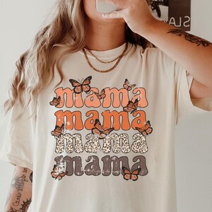 May include: A white t-shirt with a graphic print of the word "mama" repeated four times in a stacked design. Each word is in a different color and has a butterfly design around it. The shirt is a relaxed fit and has a crew neck.