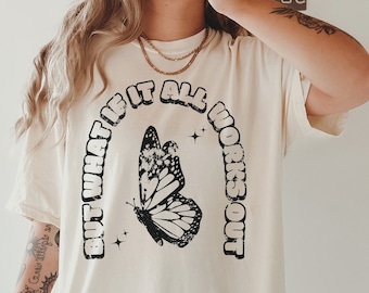 Può includere: T-shirt color avorio con grafica a farfalla nera e la scritta "BUT WHAT IF IT ALL WORKS OUT" ad arco. La maglietta ha una vestibilità ampia, adatta per un look casual.