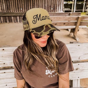 May include: A brown camouflage trucker hat with a green bill and the word "Mama" embroidered in black on the front.