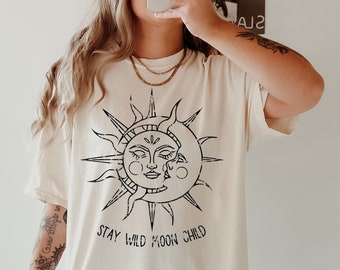 Op de afbeelding: Ivoor kleurig t-shirt met een zwarte afbeelding van een zon en maan en de tekst "Stay Wild Moon Child". Het shirt heeft korte mouwen en een losse pasvorm. Het woord "ivory" staat rechtsonder.