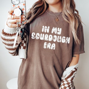 May include: A brown t-shirt with the text "In My Sourdough Era" in white, retro-style lettering. The shirt is oversized and has a relaxed fit.