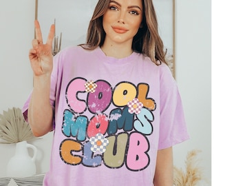 Peut inclure: Un t-shirt violet avec un graphisme vieilli qui dit "Cool Moms Club" en lettres colorées de style rétro avec des fleurs.