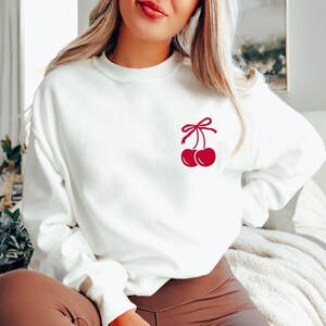 May include: White sweatshirt with a red cherry graphic on the front. The cherry has a red bow on top.
