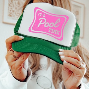 May include: Green and white trucker hat with the text "It's Pool Time" in pink. The hat has a mesh back and a curved brim. The text is in a rectangular shape.
