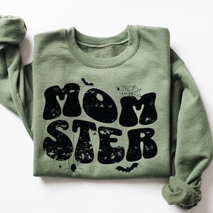 May include: A green sweatshirt with the text "MOMSTER" in black, retro-style lettering. The text is surrounded by small black spiderwebs, bats, and stars.