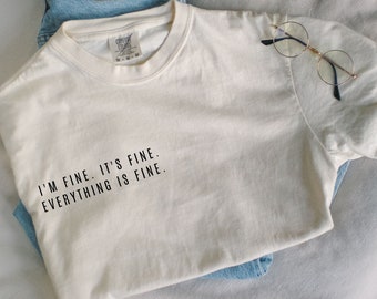 Könnte beinhalten: Ein weißes T-Shirt mit dem Text "I'm fine. It's fine. Everything is fine." auf der Vorderseite. Das Shirt liegt auf einer weißen Oberfläche mit einer Brille und einer blauen Jeans.
