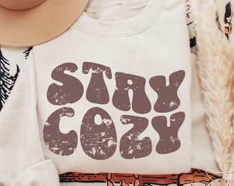 Può includere: Una felpa beige con il testo "STAY COZY" in lettere marroni in stile retrò. La felpa ha un aspetto usurato e vintage.
