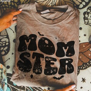 May include: A brown t-shirt with a distressed black graphic that says "MOMSTER" in a retro font. The graphic also includes a spider, a spider web, and bats.