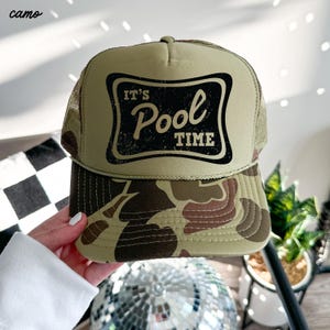 Może przedstawiać: Kremowa czapka typu trucker z daszkiem w kamuflażu i siatkowym tyłem. Z przodu czarny prostokąt z napisem "It's Pool Time" w postarzonej czcionce. Słowo "camo" znajduje się w lewym g&oacute;rnym rogu.