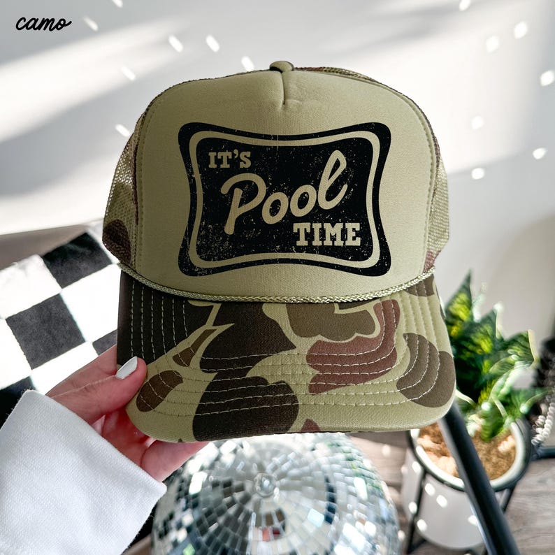 Może przedstawiać: Kremowa czapka typu trucker z daszkiem w kamuflażu i siatkowym tyłem. Z przodu czarny prostokąt z napisem "It's Pool Time" w postarzonej czcionce. Słowo "camo" znajduje się w lewym g&oacute;rnym rogu.