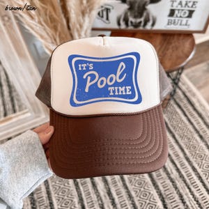 Może przedstawiać: Brązowo-beżowa czapka typu trucker z białym panelem przednim z niebieskim prostokątem z napisem "IT'S Pool TIME". Czapka ma brązowy daszek i siatkową tylną część.